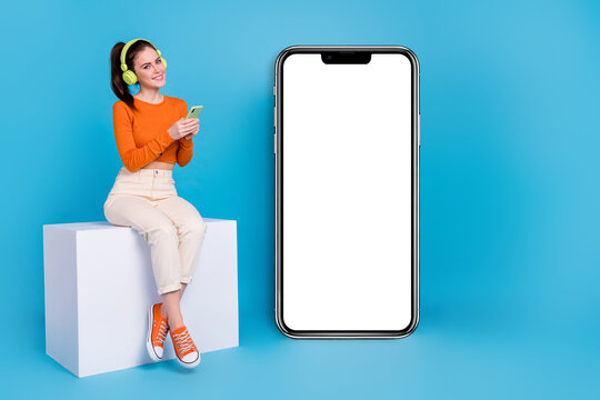 Full size photo of cute charming girl sitting podium listen her favorite song see phone promo isolated on blue color background