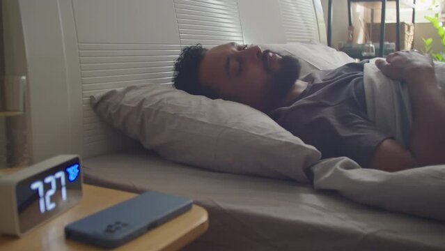 Young bearded man waking up in bed, turning off alarm clock and using smartphone in morning at home