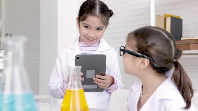 Cute elementary girls with lab coat learning chemistry in school laboratory, record exampling by notepad technology and analyzing, mixing liquid in tube in science class, education and science concept