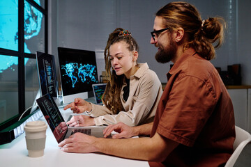 Young confident female programmer pointing at laptop screen with coded data while exlaining something to male colleague at night meeting