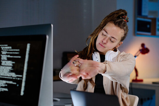 Young tired female IT support manager with dreadlocks stretching arms in front of herself while having minute of relaxation by workplace