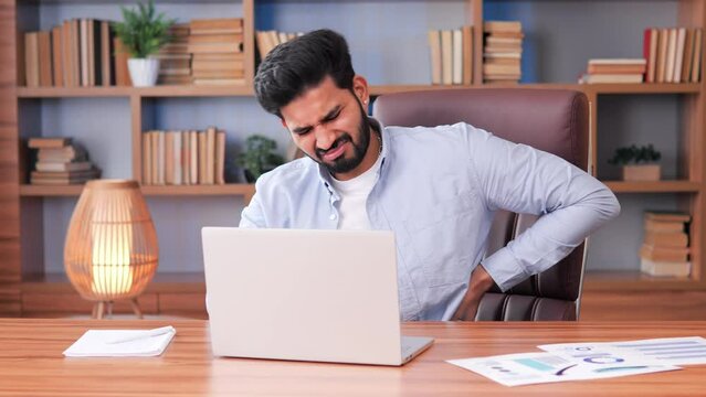 Exhausted busy Indian businessman works on his computer in the office, feeling tired and having back pain from overwork. Male man neglects his health due to long hours, stress, and pressure. 