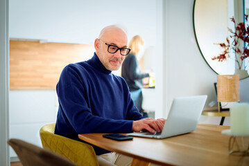 Mid aged man wearing casual clothes and using laptop for work while sitting at home