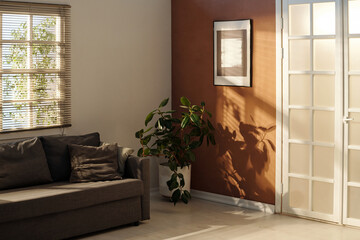 Part of spacious living room with grey comfortable couch with cushions standing by window and green domestic plant in the corner