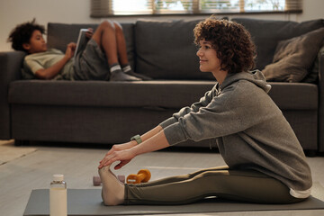 Young happy brunette woman in activewear sitting on mat on the floor of living room and stretching...