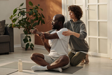 Young woman helping her husband with exercise for stretching arm muscles while sitting behind him on the floor of living room