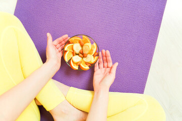 Close-up of a girl's hands, dressed in a yellow yoga suit, sitting on a yoga mat, holding a bowl of...