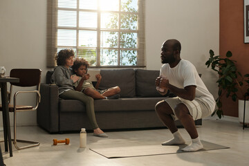 Young African American man with dumbbells squatting while standing on mat and practicing physical...