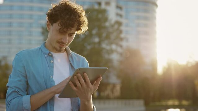 Caucasian Bearded Man Standing on the Street Surfing Internet on Tablet Computer Outdoors. Young Male Outside Using his Tablet, Texting With Friends. Male User Chatting Browsing Online