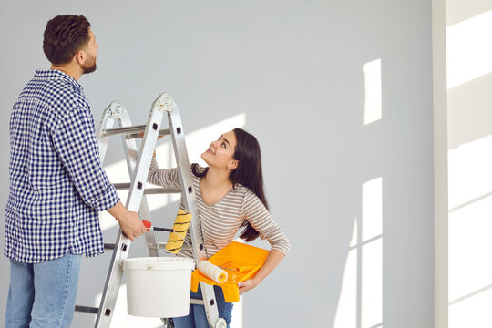 Family couple doing renovations, painting walls and whitewashing ceiling at home. Happy young man and woman standing on step ladder, holding paint can, tray, and rollers, looking up, and smiling