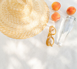 Beach vibe and still life minimalist photo. Tropical summertime picnic in the shadow. A closeup of summer accessories: sunglasses and a straw hat; a bottle of water and peaches refresh the sunny day.