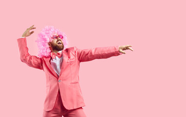 Studio portrait of happy goofy energetic bearded adult man wearing funky pink party suit, crazy curly clown wig and disco eyeglasses having fun and dancing on solid pink colour background