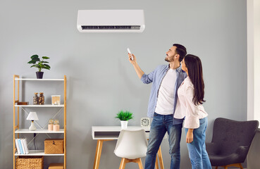 Young family couple using their modern air conditioning system at home. Happy husband and wife setting up the temperature on their white AC on the wall in the living room. Air conditioner concept - obrazy, fototapety, plakaty