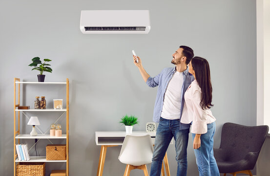 Young family couple using their modern air conditioning system at home. Happy husband and wife setting up the temperature on their white AC on the wall in the living room. Air conditioner concept