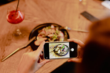 Cropped photo of woman sitting at wooden table near black plate with vegetable salad and glass with red cocktail, fork, knife in restaurant cafe, holding smartphone, taking pictures of food. Top view.