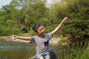 Asian boy taking a break from playing in a stream Sit on the rock, smiling brightly, arms outstretched to enjoy the splashing water happily. soft and selective focus.