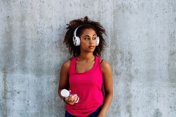 Young multiracial girl in sportswear resting after jogging in city, drinking water from sustainable bottle and listening music in headphones. - Powered by Adobe