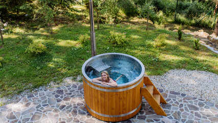 Young woman enjoying wooden bathtub with a fireplace to burn wood and heat water in backyard in mountains. High angle view.