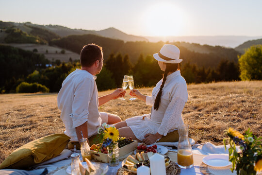 Attractive couple in love enjoying picnic and drinking wine on the hill at sunset. - Powered by Adobe