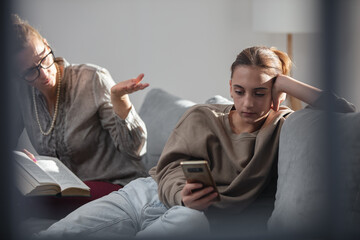 Mother having trouble with a lazy teenager girl at home while playing video games on a smartphone.