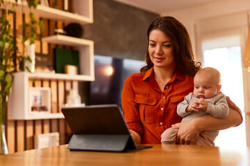 A pretty mother blogger using a digital tablet on the stand while holding her baby.