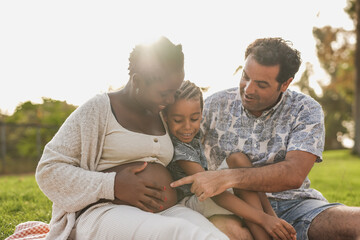 Fototapeta premium Multiracial family touching belly outdoor - Pregnant african mother, caucasian father and mixed race son having tender moment at city park - Focus on belly