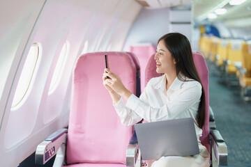 Young Asian attractive business woman passenger sitting on business class luxury plane while using smart phone mobile to take a photo and use computer laptop during the flight