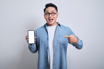 Smiling asian young man wearing denim shirt pointing to mobile phone while showing blank empty screen