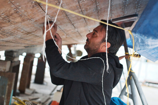 A middle-aged or young man working on the hull of a boat - putting cotton caulk into the cracks between wooden planks (caulking)