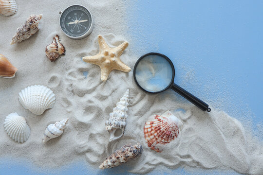Magnifier, compass, sand and seashells on blue background, research concept