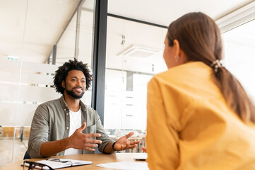 Two happy diverse colleagues African American man and Caucasian woman looking at each other and sitting in office lobby hall, man is gesturing hands