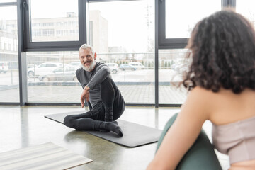 Cheerful mature coach looking at blurred woman while sitting on mat in yoga studio.