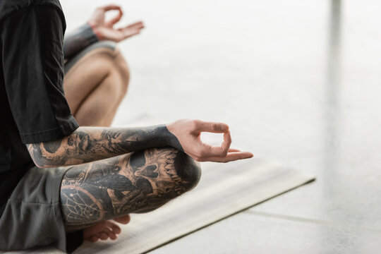 Cropped view of young tattooed man practicing gyan mudra on mat in yoga studio.
