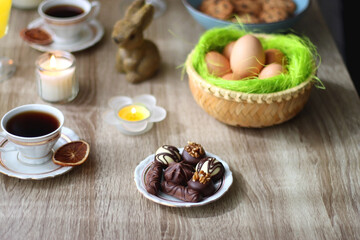 Easter eggs in the basket, bowl of cookies, chocolate pralines, Easter bunny figurine, cups of tea, glasses of juice, flowers and lit candles on the table. Selective focus.
