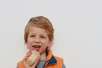 A blond boy licks an ice cream against a white wall. Happy five year old boy.