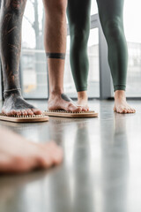 cropped view of tattooed man standing with bare feet on nail board near woman in yoga studio.