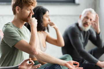 Redhead man practicing nostril breathing near blurred coach in yoga studio.