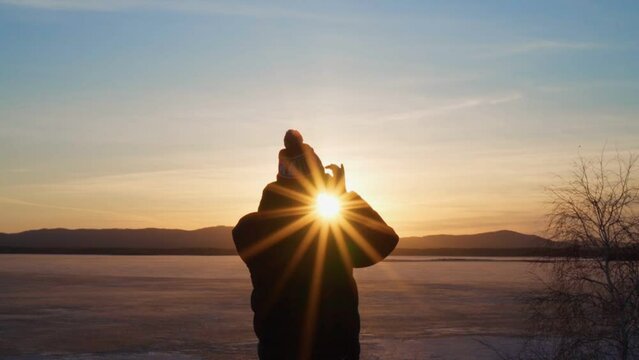 A young man looks through binoculars at a frozen lake and mountains at sunset. The rays of the bright sun gild everything around. Travel and tourism.