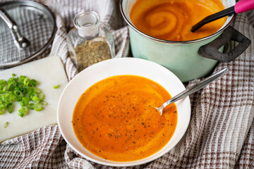 Soup from baked pepper and tomato in plate and pot, lid, herb and shoot on table.