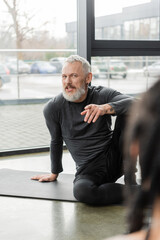 Smiling mature coach pointing at blurred african american woman in yoga class.