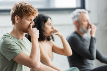 Redhead man practicing nostril breathing near blurred group in yoga studio.