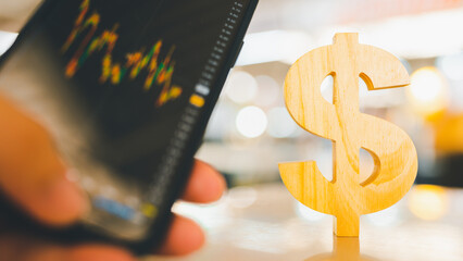 Model of a wooden dollar sign on the table, together with a businessman's hand looking at a stock...