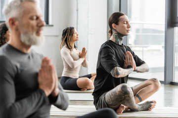 Tattooed man practicing anjali mudra near multiethnic people in yoga group.