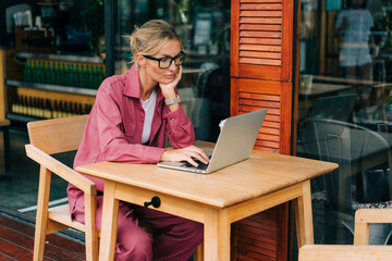 Business woman in glasses sitting at a table in a cafe uses a laptop for remote work.