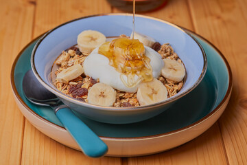 Bowl of granola with banana,yogurt with honey being drizzled over it.