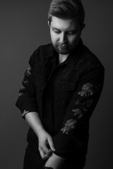 Black and white studio portrait of a handsome male model wearing a black jacket with embroider roses on it. He is caucasian with light hair and he is in his 20s. 
