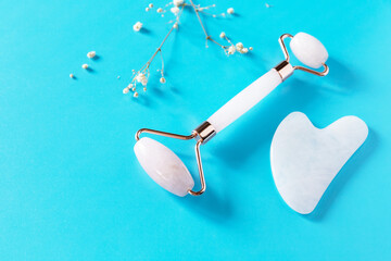 Rose quartz facial massage roller with gypsophila flowers over blue background. Massage tools with...