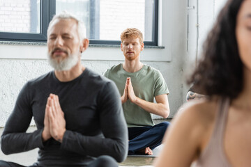 Redhead man meditating with anjali mudra near blurred people in yoga class.