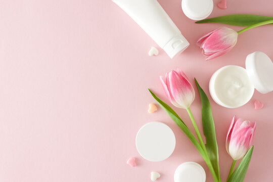 Organic skincare concept. Top view photo of cosmetic tubes without label open cream jar colorful hearts and pink tulips on pastel pink background with empty space