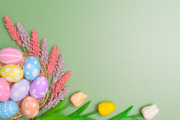 Happy Easter holiday greeting card design concept. Colorful Easter Eggs and spring flowers on pastel green background. Flat lay, top view, copy space.
